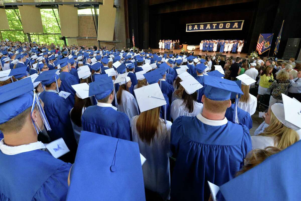 Students wearing white and blue graduation caps face the auditorium stage